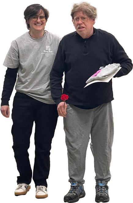 Dos personas, una más joven y otra más mayor, caminan juntas hacia delante. Uno de ellos sostiene una flor y unos papeles.