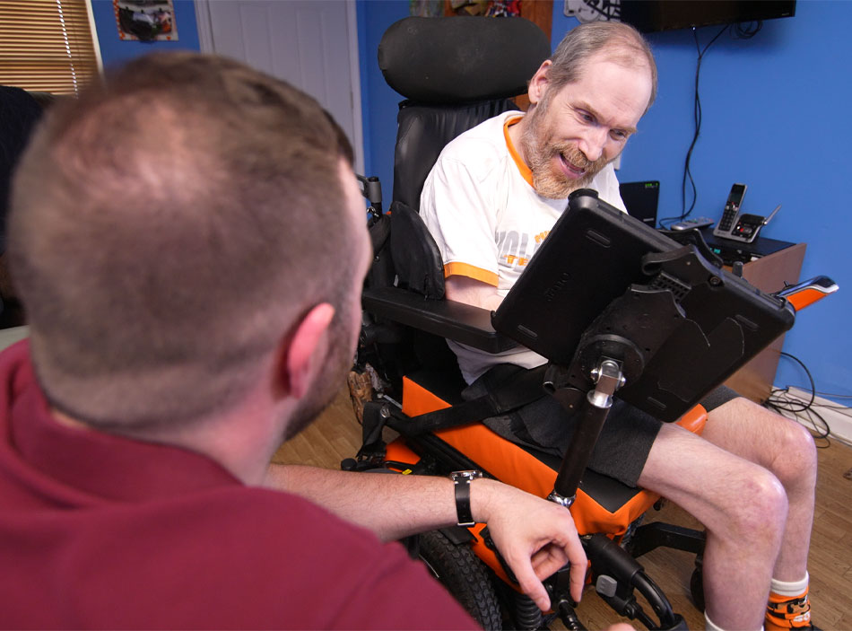 A man with short hair and a beard wearing a t-shirt and shorts looks down at a computer screen on a wheelchair he is sitting in.