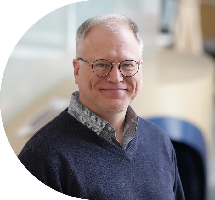 Un hombre con el cabello corto y gris y gafas sonríe levemente mientras mira al frente.