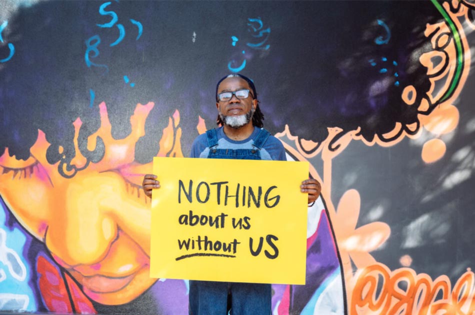A man with glasses holds a “Nothing About Us Without Us” sign as he stands in front of a mural.