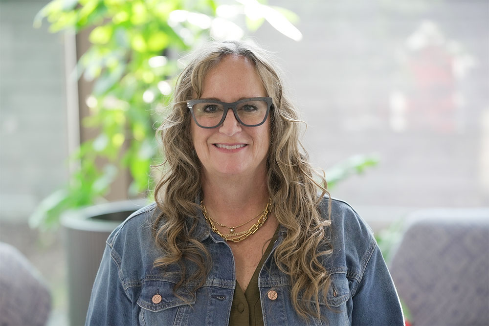 A woman with long, wavy blonde hair and glasses is smiling at the camera. She is wearing a denim jacket over a dark t-shirt that has a white graphic design and the words 'Lucky Trails - Arizona.' The background features a modern indoor setting with soft, neutral colors and some colorful furniture, suggesting a casual and welcoming environment.