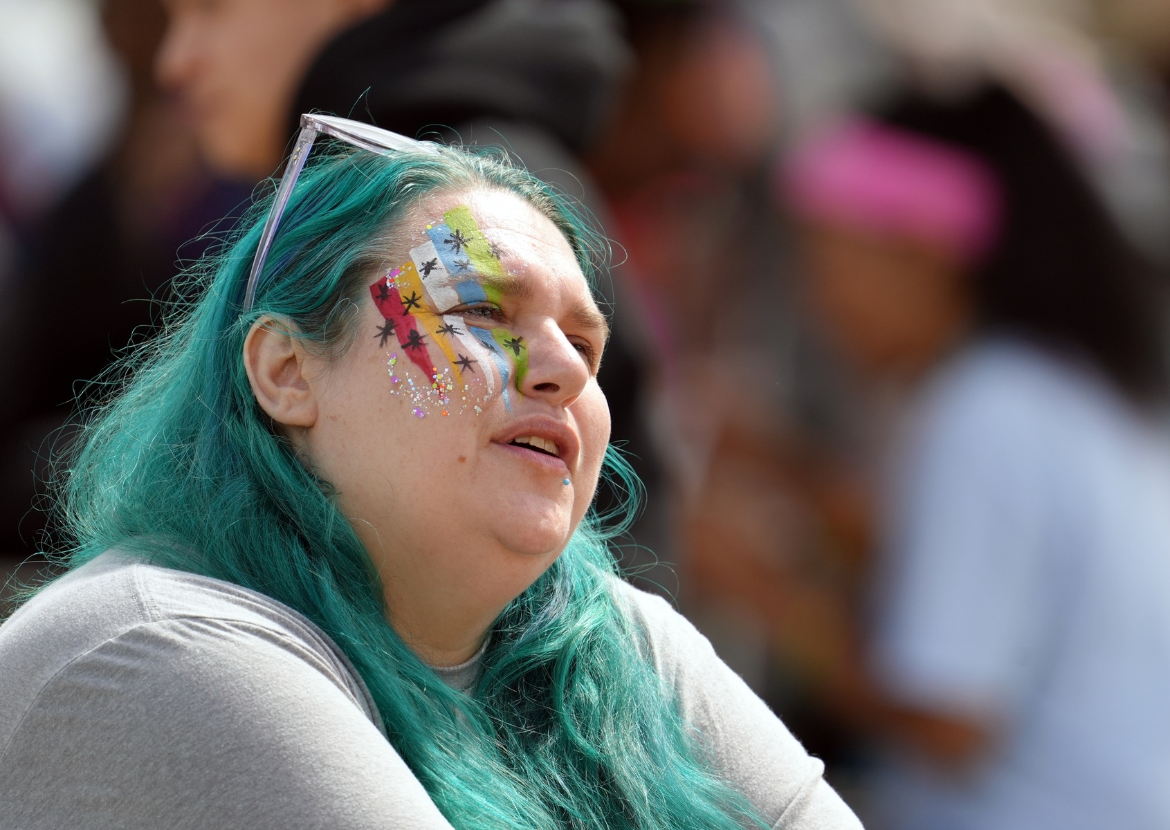 A woman with aqua-blue hair and painted symbols on her face looks ahead.