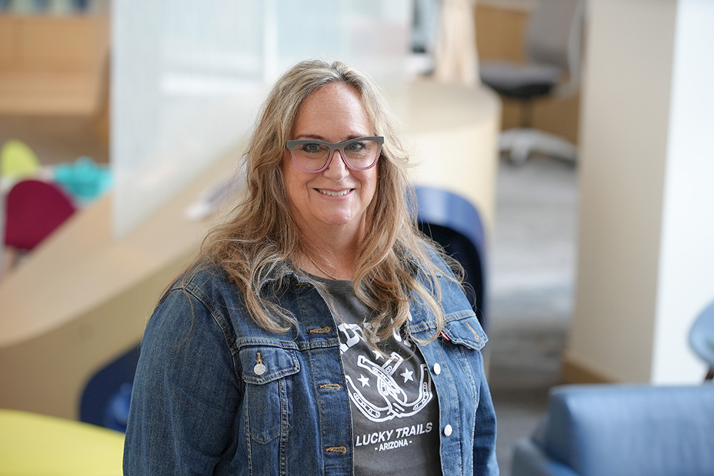 A woman with long, wavy blonde hair and glasses is smiling at the camera. She is wearing a denim jacket over a dark t-shirt that has a white graphic design and the words 'Lucky Trails - Arizona.' The background features a modern indoor setting with soft, neutral colors and some colorful furniture, suggesting a casual and welcoming environment.