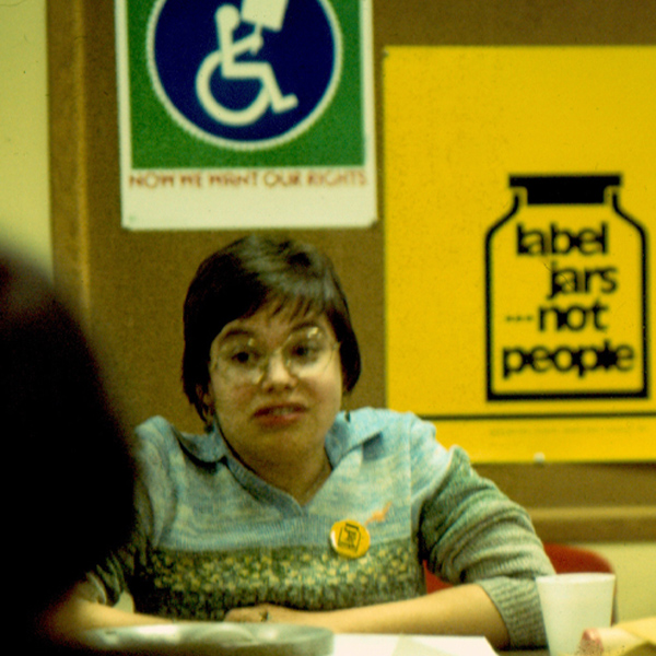 Woman seated at table with posters behind her reads Now Give Us Our Rights and Label Jars ... Not People.