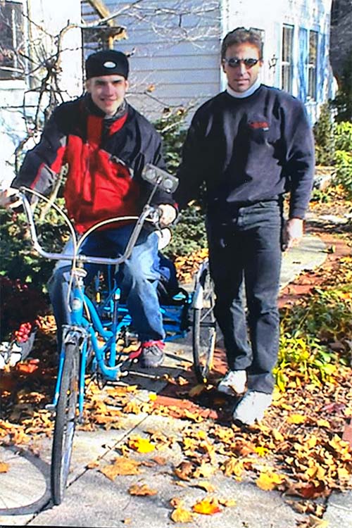 Two men are outside a house on a spring day. Renzo is on a blue three-wheel bike. He is wearing jeans, a red and black jacket, and has a black baseball hat on backwards. The other person is standing beside him. They are both looking straight at the camera. He is wearing black jeans and a sweatshirt with a white turtleneck and sunglasses.
