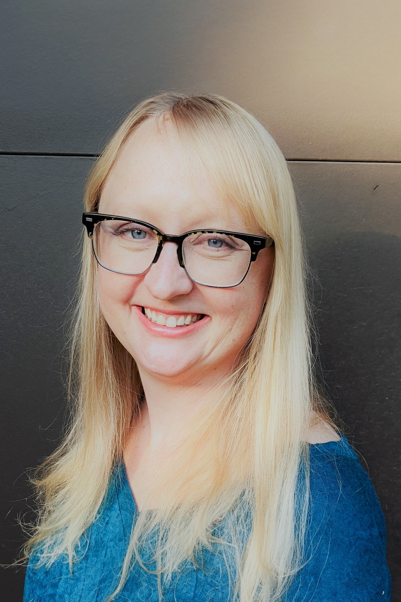 A woman with light skin and blonde hair is wearing glasses, a light gray blazer, and a light pink top. She is smiling, and the background features blurred colorful elements that resemble artwork or decorations.