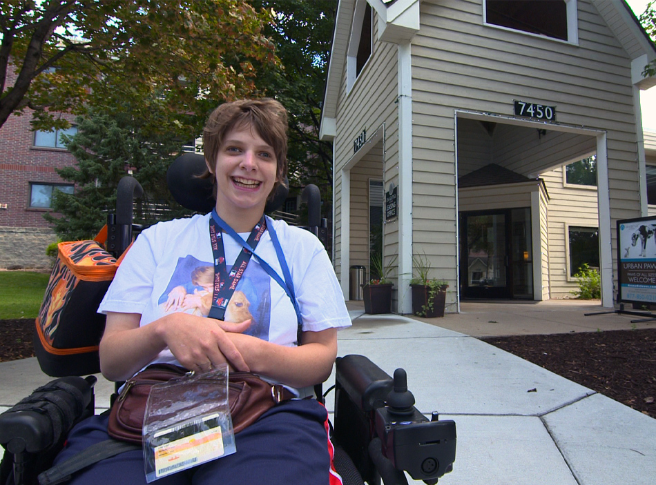 A woman in a wheelchair wearing a t-shirt with credentials around her neck smiles.