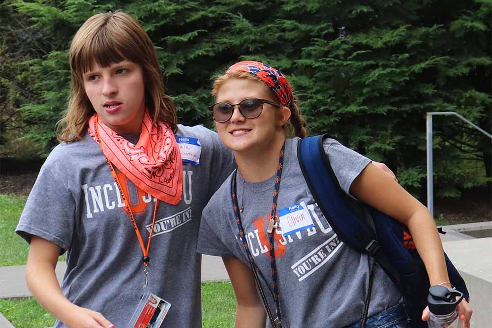 Two young, female college students. They are outside and are wearing name tags. Their t-shirts read "Inclusive U." One of the students has an intellectual disability.