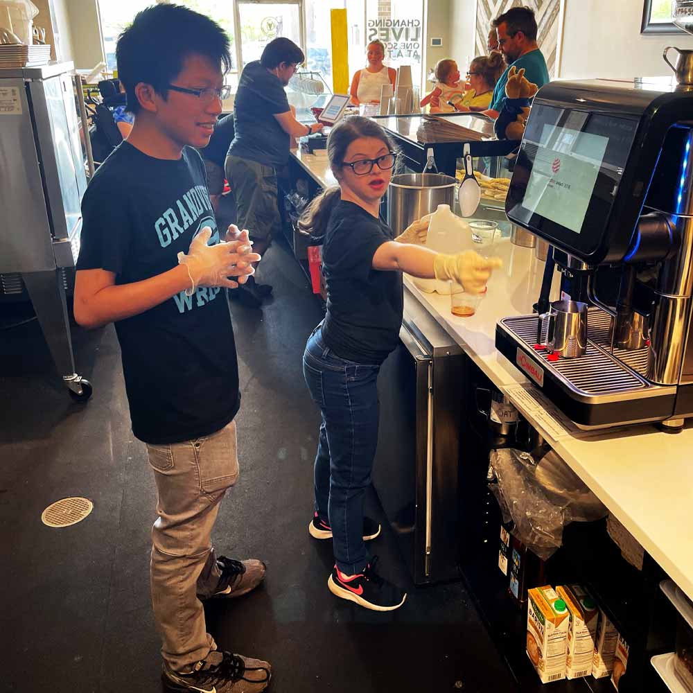 Unos jóvenes trabajan detrás del mostrador de una heladería. Un joven con camiseta negra y pantalones color caqui junta las manos mientras utiliza unos guantes. Una mujer joven con pantalones vaqueros, gafas y coleta, también con guantes, señala un dispensador de bebidas.