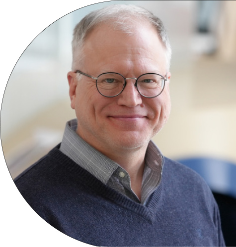 A man wearing a blue sweater and over a collared shirt looks at the camera with a slight smile.