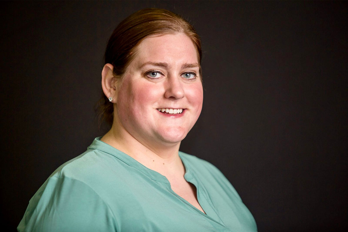 Mary is smiling while looking at the camera. She is standing with her right shoulder forward and left shoulder behind. She has longer reddish-brown hair that is pulled back, green eyes and is wearing diamond earrings and a light green V-neck top.