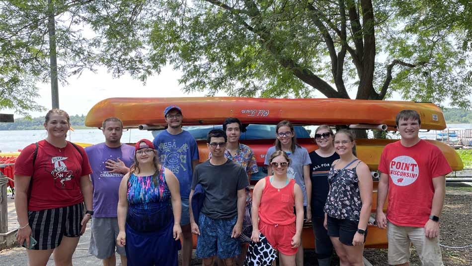 Un grupo de exalumnos universitarios con shorts y camisetas se paran frente a los kayaks cerca de un lago.