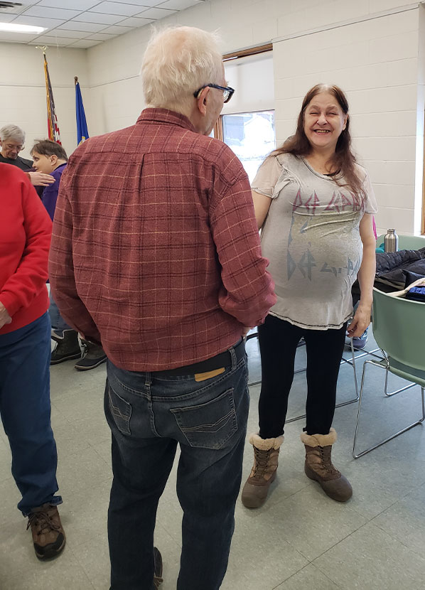 A man standing with his back o the camera and is facing Paige who is smiling. They visiting with each other are at the Community Center. Page is smiling, has long brown and white hair, and is wearing a Def Leppard top, black leggings and snow boots.