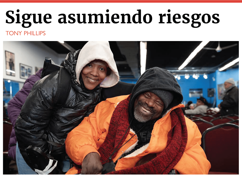 Una mujer y un hombre sonrientes aparecen en una habitación con una cálida iluminación, con filas de sillas rojas y varias personas al fondo. La mujer está de pie e inclinada hacia delante, lleva una chaqueta con capucha de color claro y un abrigo negro. El hombre está sentado, lleva una sudadera oscura con capucha, una bufanda roja y una chaqueta naranja brillante. Ambos parecen felices y cómodos, y el hombre muestra una amplia y alegre sonrisa. El titular que aparece encima dice «Retirarse con un propósito» y la firma es de Tony Phillips. El escenario sugiere un evento comunitario o social, posiblemente en un espacio recreativo o de reunión.