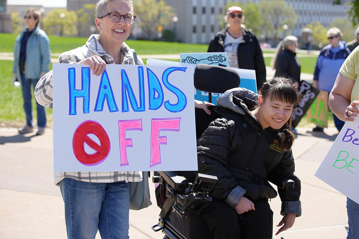 Standing on the right is Cindy, with short gray hair, glasses, smiling, and holding a sign that says HANDS OFF. She is wearing blue jeans and a cream and gray flannel jacket. Eve is on the right. She has long dark brown hair and is smiling. She is wearing a black and gray puffy jacket and is a wheelchair user. They are outside on a sunny say among other people who are also holding signs at the rally.