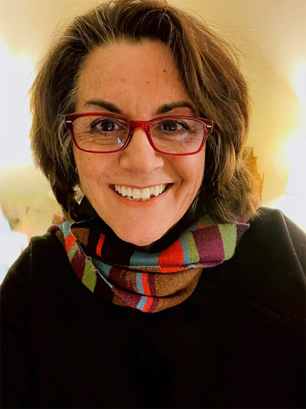 LeeAnne Kingsbury is smiling while looking at the camera. She has shoulder length brown hair; red glasses She is wearing a black top with a colorful stripped scarf.
