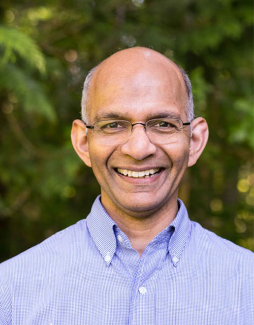 A balding man with some white and gray hair on the sides of his head, glasses, big smile, blue oxford button down shirt, with trees in background.