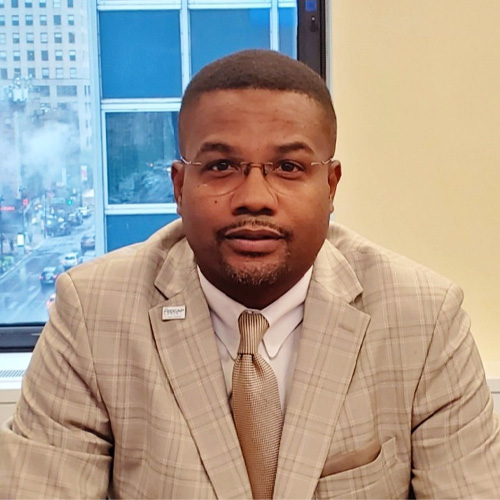 Man, short black hair, mustache and beard around mouth, glasses, light brown plaid suit, with cream colored shirt and light brown tie.