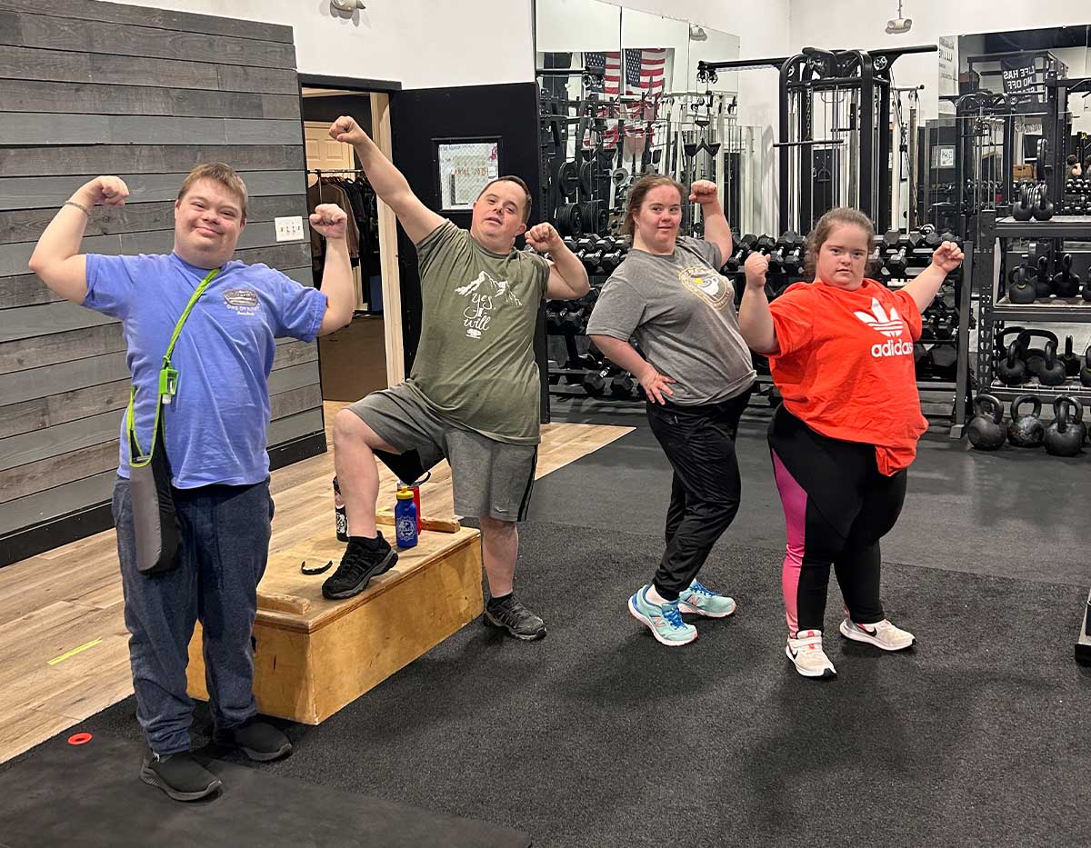 Cuatro personas —Charles, Chris, Lauralee y Briley— están de pie, uno al lado del otro, flexionando los brazos y sonriendo tras completar un entrenamiento en I CAN.