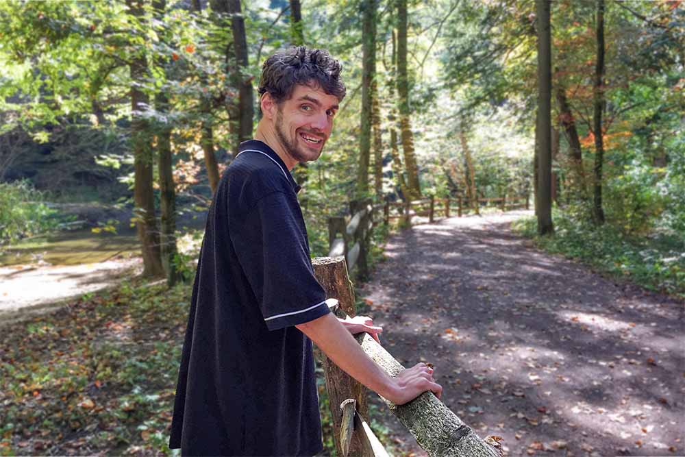 A man with light skin and curly brown hair is standing outdoors on a dirt path surrounded by a lush forest. He is wearing a black shirt with white trim and is leaning on a wooden fence, smiling at the camera. The path is lined with trees, and sunlight filters through the foliage, creating a serene and natural setting. He is standing with his hands on a fence.