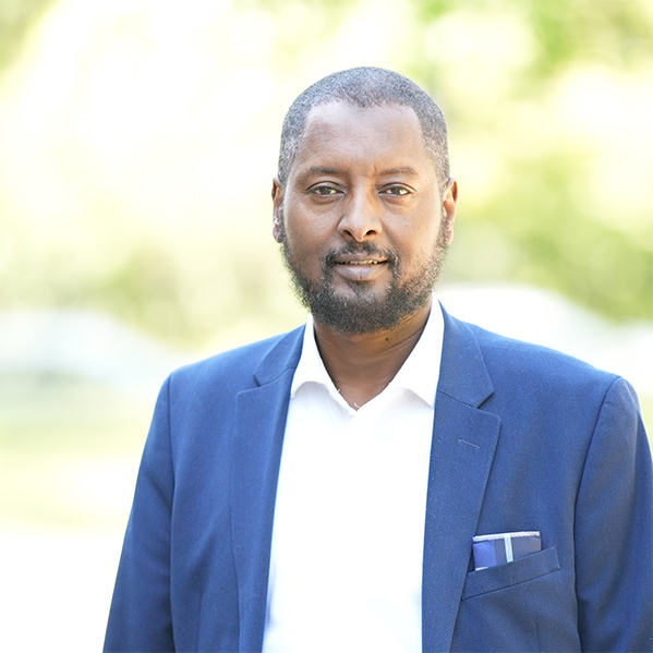 Mahdi Warsama is outside in front of green trees. He has a slight smile as he looks at the camera. He has short black hair and beard. He is wearing a blue sports coat over a white oxford. He has a blue checked pocket square.