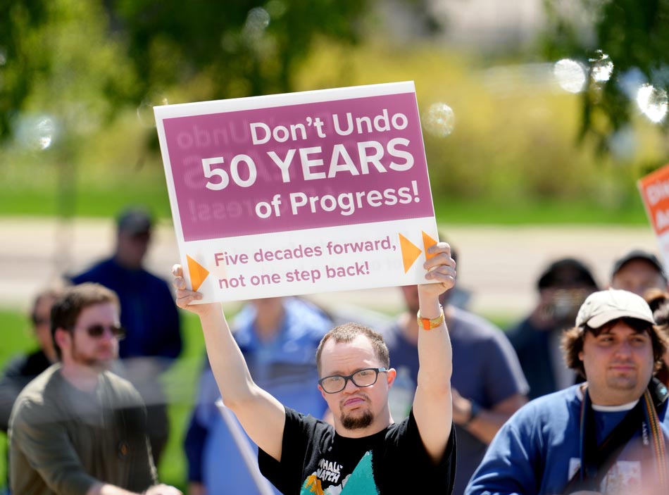 A group of people at a rally. One man in a black t-shirt holds up a sign that says “Don’t Undo Years of Progress!”