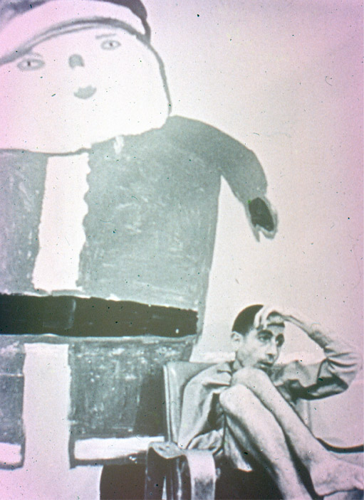 A very thin man sits in a chair with his knees to his chest. Behind him is a large primitive drawing of a Santa-like character.