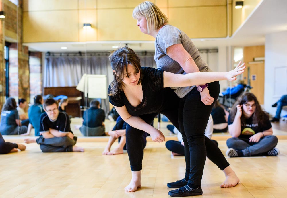 Two white women, one with dark hair and the other blonde, demonstrate a move to seated students in the background. The woman with dark hair is bent over, resting her right elbow on her knee while her left arm and leg extend. The woman with blonder hair stands in profile, legs straight, slightly bending at the waist into the side of the other woman.