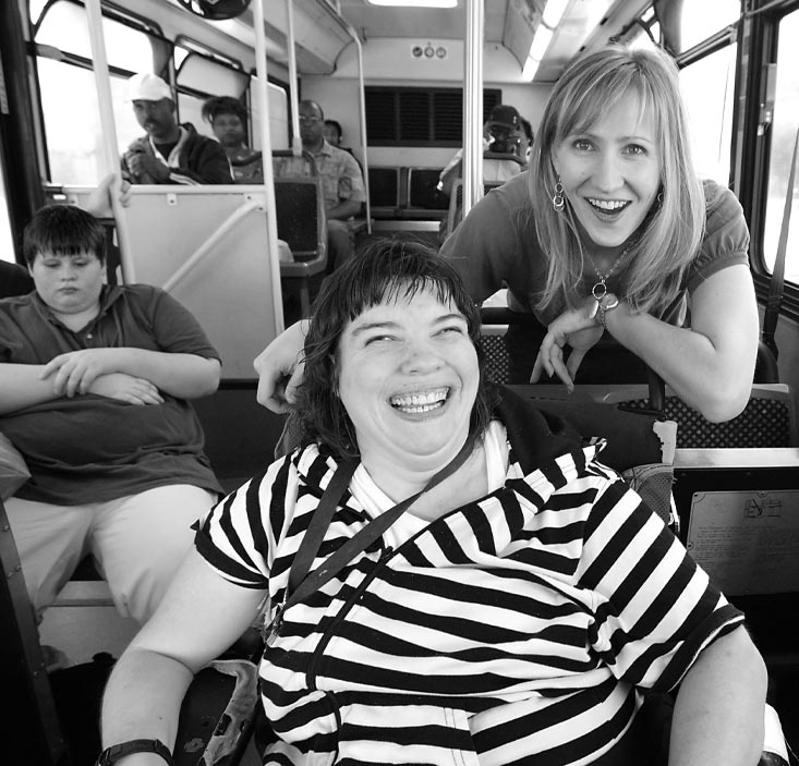 Several people ride a train. Two women in the foreground smile while looking at the camera.