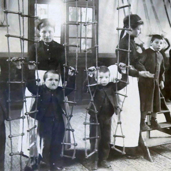 Niños pequeños en un aula de la década de 1870 sosteniendo escaleras de cuerda mientras los profesores los observan.