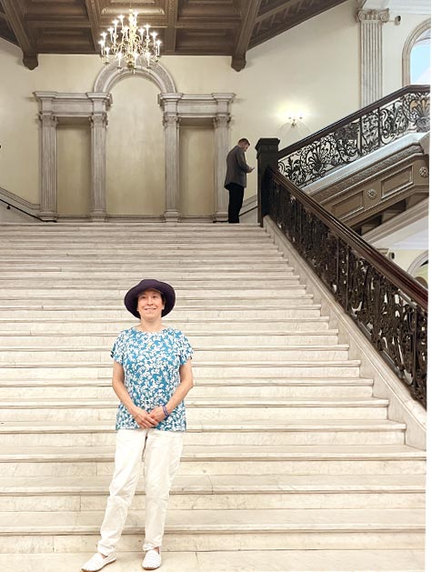 Una mujer con sombrero de ala, camisa azul estampada, pantalones y zapatos blancos permanece de pie con las manos juntas en el primer peldaño de una escalera de mármol de un edificio gubernamental.