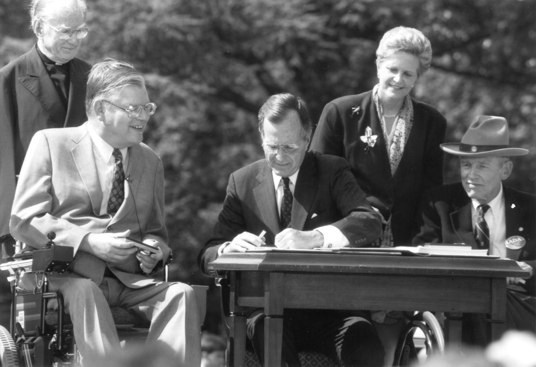 Two seated men flank President Bush, also seated, as he signs an important document. Another woman and man stand behind them.