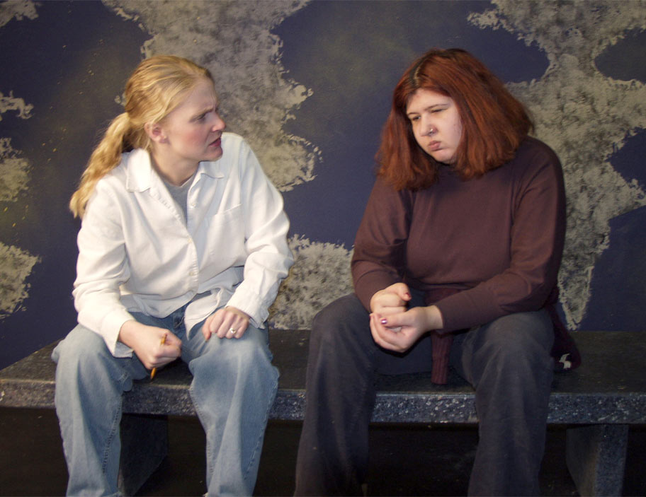 A blonde woman in a white jacket looks seriously at a woman sitting next to her. The other woman looks away, appearing sad.