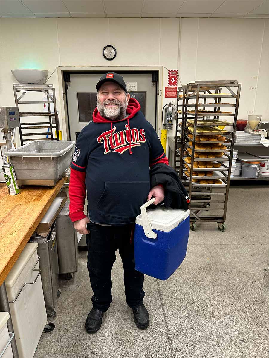 Jim is standing the kitchen of the bakery at the store he works at. Behind him are racks with bakery items, beside him is work counter, with a large bin, under the counter are appliances. Jim has a big smile and is looking at the camera. He has a gray and white beard, is wearing a Minnesota baseball hat and sweatshirt, black pants and shoes. He is holding a dark blue and white lunch cooler in his left hand.