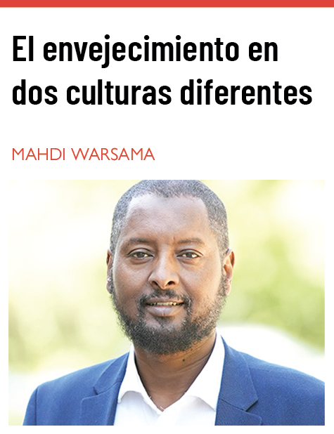 Un hombre con la barba bien recortada y el cabello corto está de pie al aire libre. Lleva una chaqueta azul sobre una camisa blanca y mira directamente a la cámara con una expresión amable y segura. El fondo está suavemente difuminado con luz natural y vegetación, creando un ambiente cálido y acogedor. El texto que aparece sobre la imagen dice: «El envejecimiento en dos culturas diferentes: «Cuidado de personas mayores somalíes en Minnesota», artículo escrito por Mahdi Warsama.