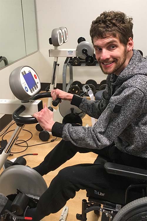 Renzo is working out at the gym on equipment that he uses his feet to peddle while in his own wheelchair. Renzo is wearing a black and gray speckled hooded sweatshirt and black sweatpants. He has short dark brown hair, and mustache and beard.