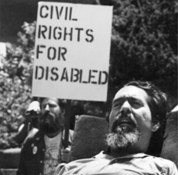 A man with a goatee sits in a wheelchair while another man behind him holds a sign reading Civil Rights For Disabled.