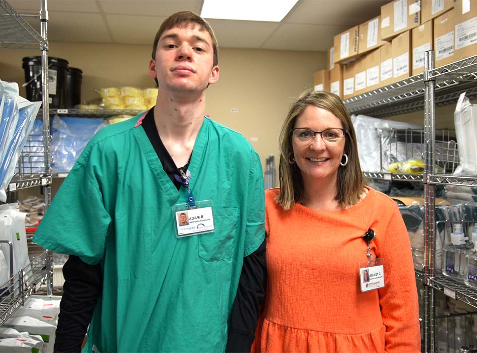 Un joven alto vestido con ropa de hospital y una tarjeta de identificación está de pie junto a una mujer que lleva una camiseta naranja y una tarjeta de identificación.