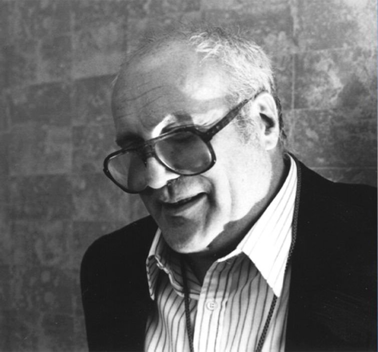 Un hombre con gafas rectangulares grandes y cabello gris, vestido con camisa con cuello y chaqueta de traje.