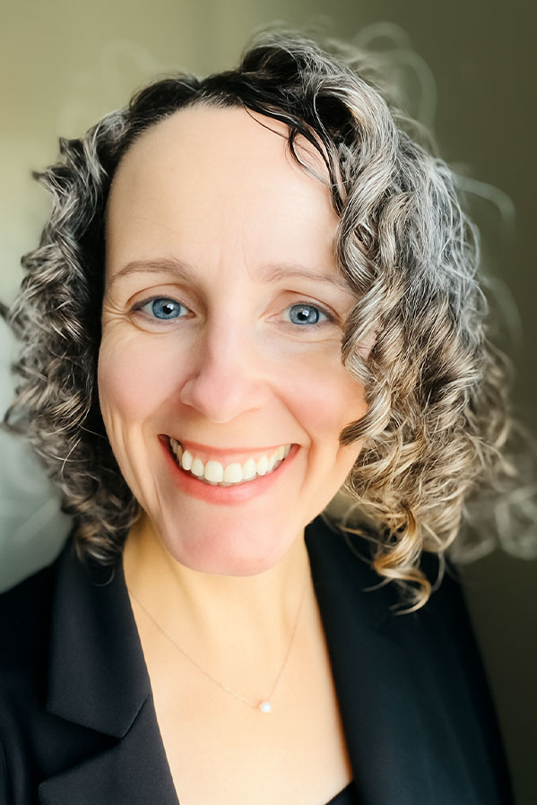 Bethany is smiling at the camera. She has curly brown, grey and white hair, blue eyes and is wearing a black top and jacket along with a thin gold chair with a single small pearl.