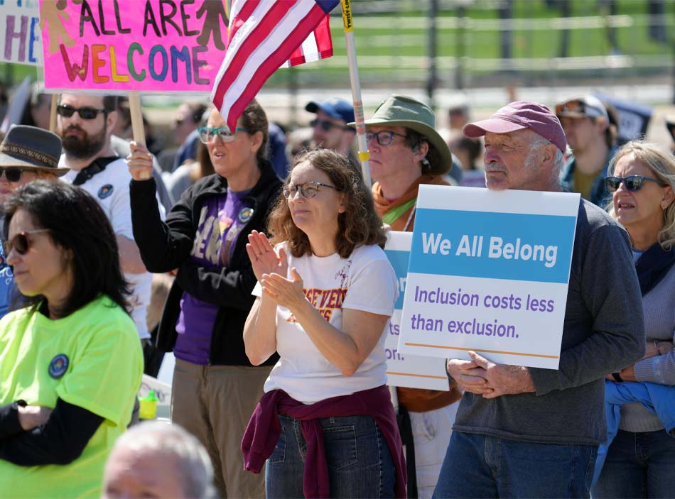 Los manifestantes, que llevan una bandera y pancartas sobre la inclusión y la pertenencia, aplauden y escuchan a un orador que no aparece en cámara.