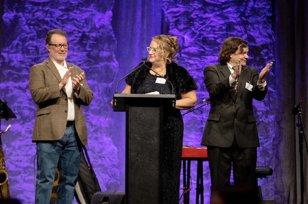 Brad Perry, Amy Hewitt, and Nathan Perry onstage at The Arc Minnesota's Gala. Amy Hewitt is at the podium.