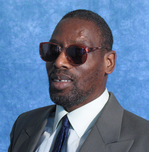 Man with dark sunglasses, short dark hair, and beard, wearing a suit with a necktie.