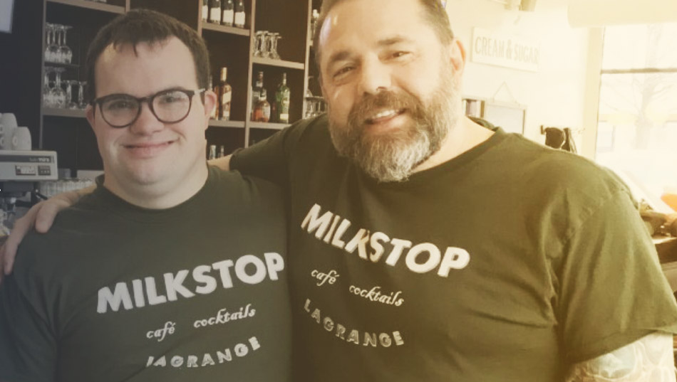 Two male coworkers wearing identical t-shirts stand with arms around each other in a café. One man wears dark glasses, the other has facial hair.