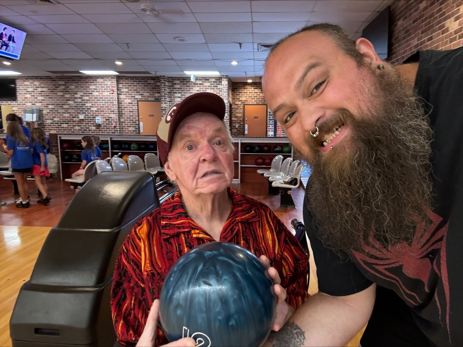 Two men in a bowling alley. Wayne is the man on the left. He has on a red, orange and yellow flame patterned shirt, and is wearing a maroon and tan baseball hat. He has white hair and blue eyes. He is holding a blue bowling ball. Tom is the nan on the right. Tom is smiling, has short dark brown hair, a nose ring and a long dark brown and white beard. He is wearing a black T-shirt with a big red spider on the front.