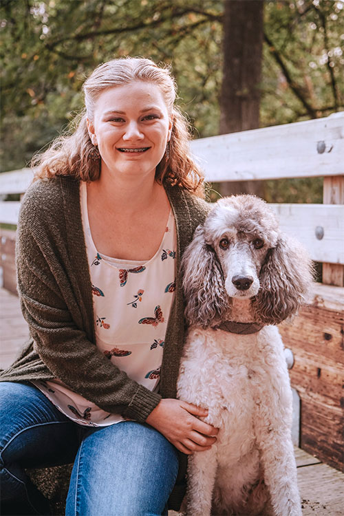 Woman with blonde hair in half ponytail wearing a moss green sweater and a shirt with butterflies. She is looking at the camera, smiling with braces, and is sitting next to a silver beige Standard Poodle.