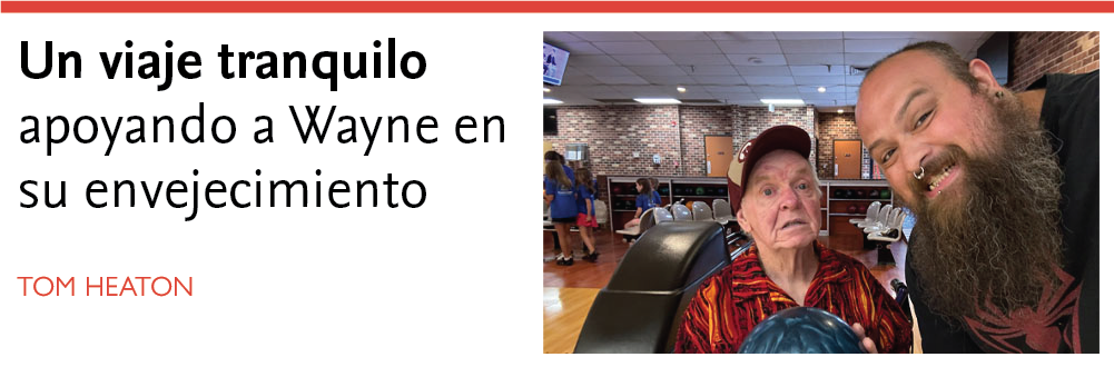 Un hombre mayor con una camisa estampada en rojo y naranja y una gorra de béisbol sostiene una bola de bolos azul en una bolera. A su lado, un hombre más joven con una larga barba, un aro en la nariz y piercings faciales sonríe ampliamente a la cámara. El fondo muestra a varias personas, entre ellas niños con camisetas azules, sentadas y de pie cerca de las pistas de bolos. El ambiente es animado y agradable. El título que aparece encima de la imagen dice: «Un viaje tranquilo: apoyando a Wayne en su envejecimiento, con Tom Heaton como autor.