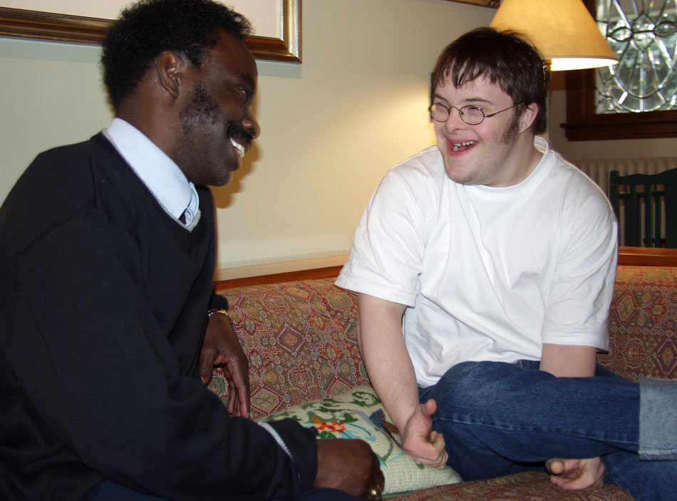 Two young men sit on a couch, smiling at each other.