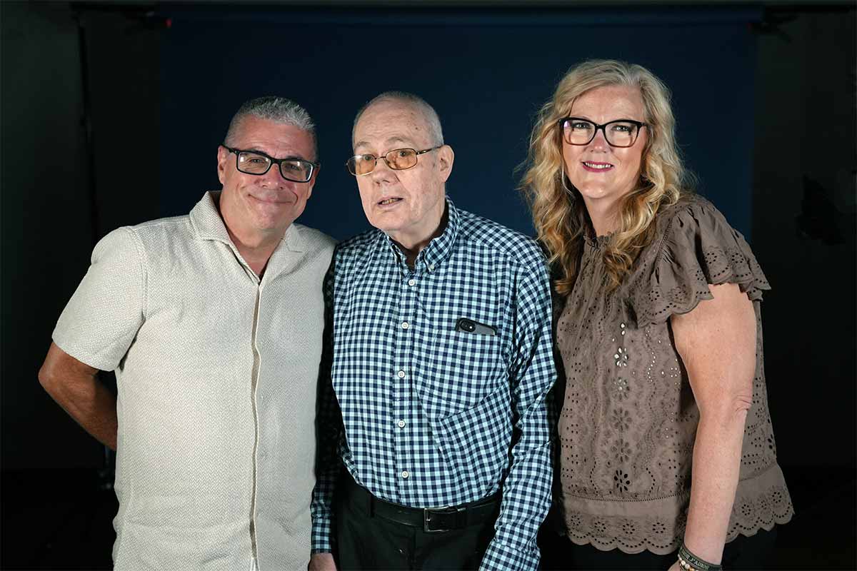 John Leo, David Liscombe and Michelle Leo are all standing and looking at the camera. Jon is on the left. He is smiling. He has short gray hair and black glasses. He is wearing a light tan short-sleeve button-down shirt. David is in the middle. He has short white hair, glasses, and is wearing a black and white checked button-down, long-sleeve shirt with black pants. He has cell phone in his shirt pocket. Michelle is on the right. She has long blondish-brown hair, glasses and is wearing a short-sleeved, chocolate colored blouse with black pants.