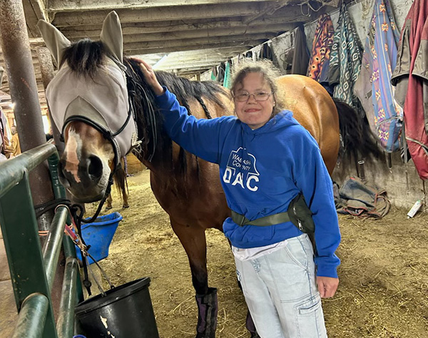 A Wabasha DAC service recipient is in the horse stable with a horse that she works with in equine therapy. She is wearing a Wabasha County DAC hoodie, and the horse is blindfolded.